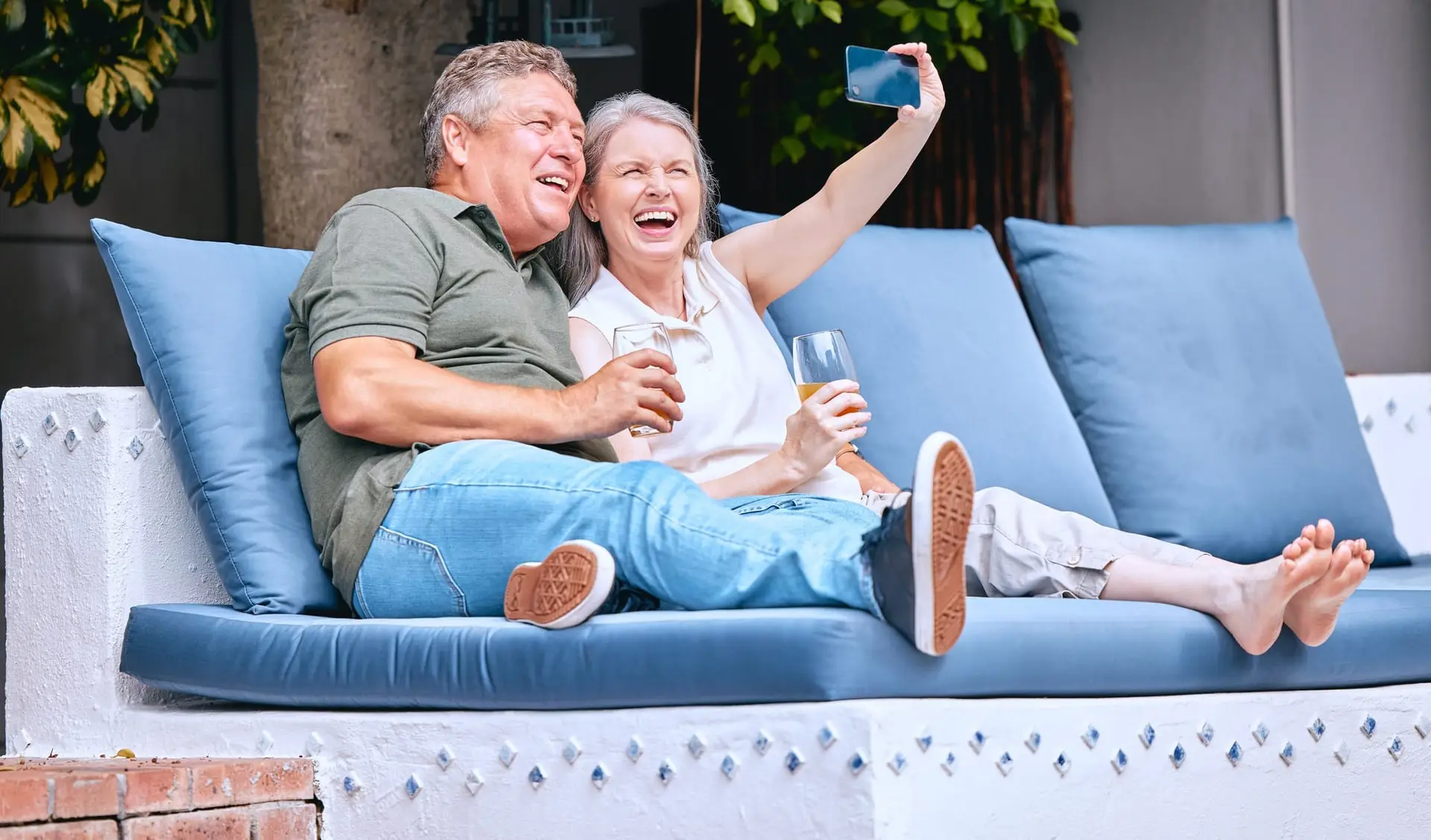 A happy senior expat couple taking a selfie while relaxing on an outdoor sofa with drinks, enjoying retirement in Spain with peace of mind from 247 Expat Insurance.